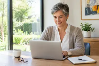 Fifty-something woman with short salt-and-pepper hair working at a laptop in a bright, modern home office with a large window, notebook, coffee mug, and minimal decor.